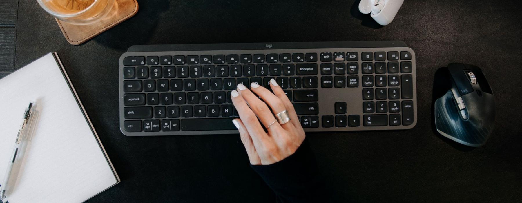 woman's hand on a keyboard surrounded by office items and a cup of tea on a coaster